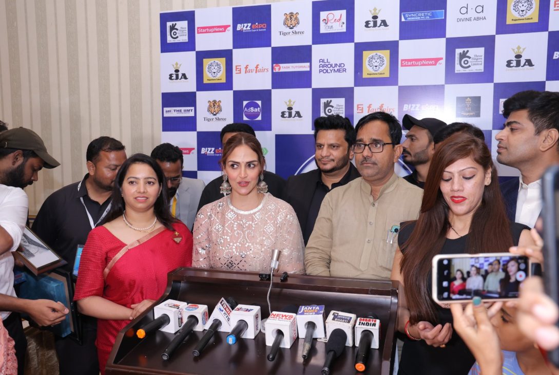 A group of people, including a woman in a red saree and a woman in a white lace outfit, standing in front of a backdrop filled with various brand logos. They are surrounded by microphones from different news channels, indicating a press event at Bizz Expo & Summit.