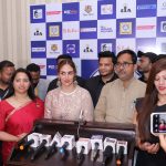 A group of people, including a woman in a red saree and a woman in a white lace outfit, standing in front of a backdrop filled with various brand logos. They are surrounded by microphones from different news channels, indicating a press event at Bizz Expo & Summit.