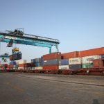 A large container ship being loaded with cargo containers at a Gujarat Pipavav Port, with cranes and railway tracks visible.