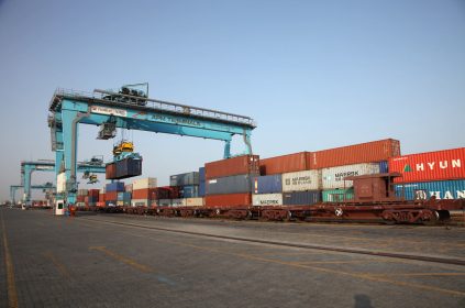 A large container ship being loaded with cargo containers at a Gujarat Pipavav Port, with cranes and railway tracks visible.